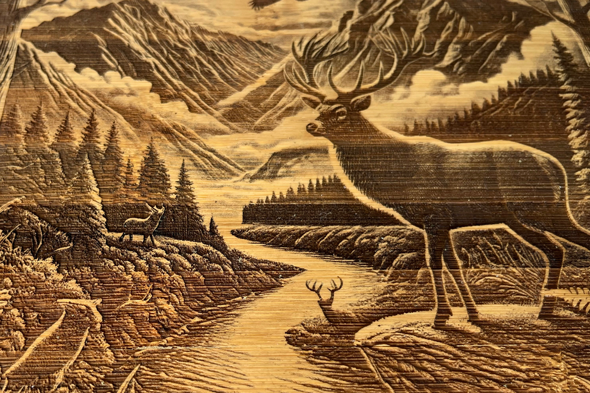 Close-up of the Hunter’s Sunrise wooden board engraving, showing the stag, mountain valley, river, surrounding forest and natural wood grain.