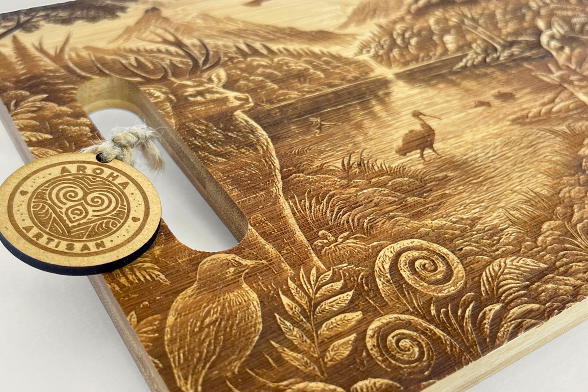Angled close-up of the Where We Belong wooden board, showing the Aroha tag, handle slot, bird and fern engraving and natural wood grain.